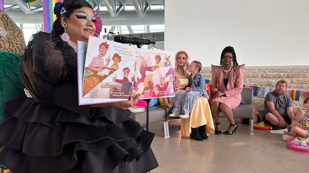 Une drag queen montre des pages de livre pour enfants dans un local où on aperçoit des gens.