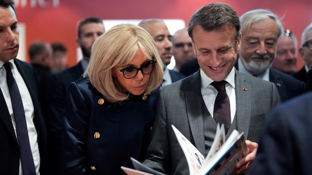Emmanuel Macron, tout sourire, et sa femme Brigitte, concentrée, feuillettent un livre dans une foule.