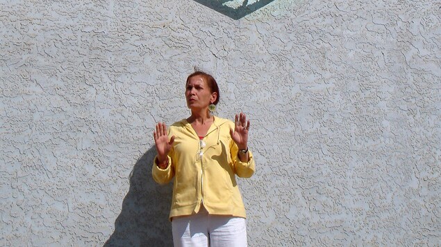 Eveline Hamon, vêtue de jaune, présente ses mains à l'appareil-photo, sous une large sculpture de fer de Joe Fafard à Duck Lake qui représente un couple franco-métis dans un cœur.