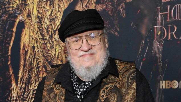 Un homme barbu portant des lunettes et une casquette prend la pose devant l'affiche de la série «Game of Thrones». 