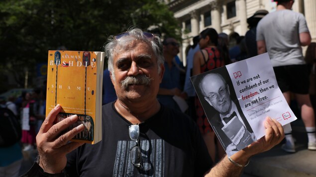 Hasan Mujtaba tient un livre et une petite affiche sur les marches d'une grande bibliothèque à New York.