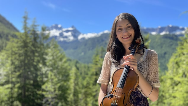 Jocelyn Pettit, souriant, tenant son violon face à la caméra. Elle prend la pose devant un paysage de montagnes.