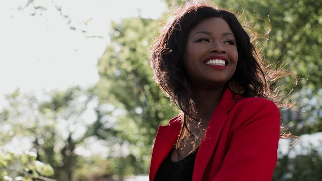 Une femme noire placée de trois-quart regarde au loin par-dessus son épaule, avec un grand sourire. Le soleil tombe sur sa chevelure lâchée au vent. Des arbres sont en arrière plan.