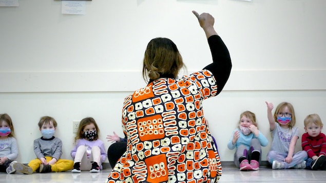 Une femme noire photographiée de dos lève le pouce en l'air. Devant elle et adossés à un mur de jeunes enfants portants des masques rient et reproduisent son geste.
