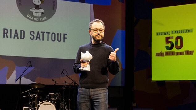 Un homme tenant un trophée en forme de chat fait un signe de la main sur scène lors d'une cérémonie de remise de prix.
