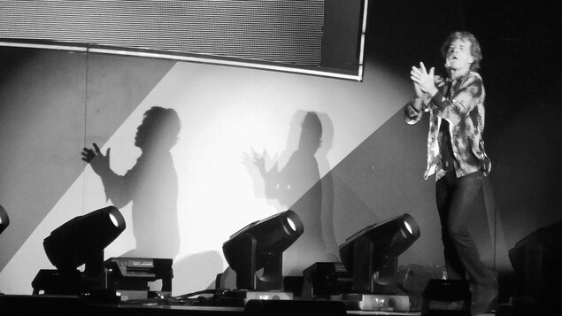 Mick Jagger sur scèene en noir et blanc, avec des ombres sur un mur derrière lui.