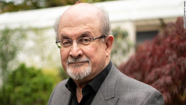 Salman Rushdie, face à l'appareil photo, en complet, devant des arbres.