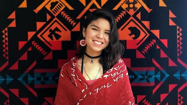 Shaunteya a des cheveux noirs de jais. Drapée dans un châle rouge, elle pose tout sourire devant une tapisserie autochtone. Elle porte aussi des boucles d'oreilles rouges au centre desquelles se trouve un cheval.