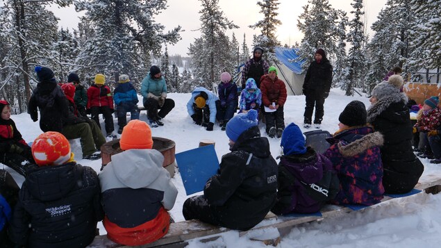 Groupe d'enfants habillés avec vêtements d'hiver assis autour d'un feu dehors avec tente et arbres recouverts de neige, le 30 novembre 2022, à Yellowknife.