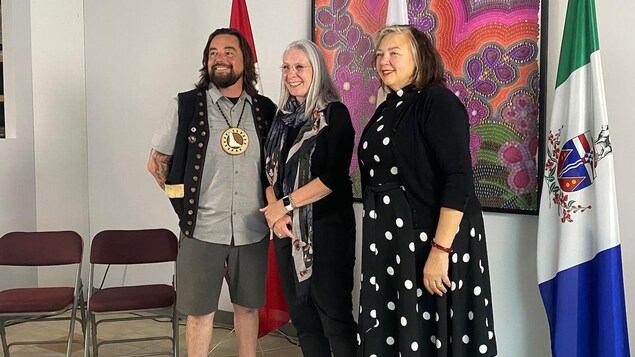 Trois personnes souriantes se tiennent devant une œuvre colorée et des drapeaux, dont celui du Canada et du Yukon.