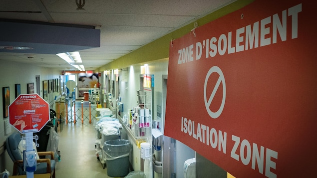 On voit en plongée un couloir d'hôpital. En gros plan, à droite de l'image, on voit un grand écriteau en rouge où il est écrit « zone d'isolement » avec le symbole d'interdiction de passer.