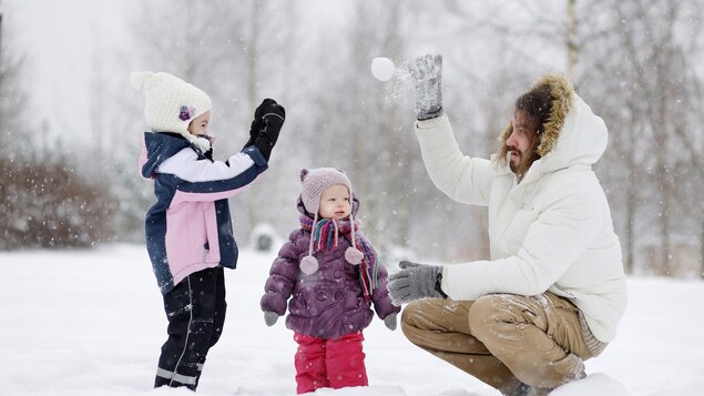 Un père et ses deux jeunes enfants jouent dans la neige et se lancent une balle de neige. 
