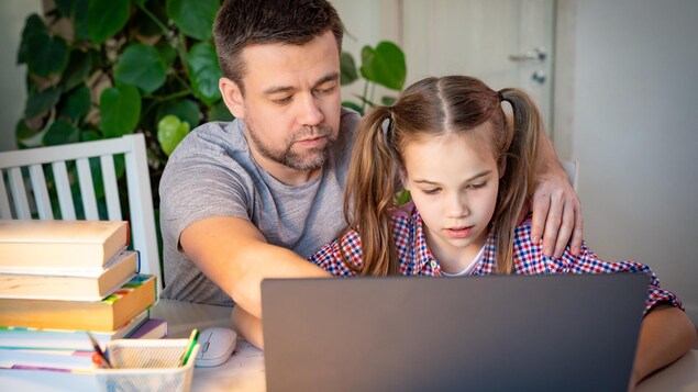 Un père et sa fille sont assis à une table, à la maison, devant un ordinateur. 