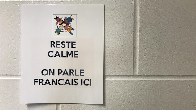 Une affiche blanche sur un mur blanc où il y est écrit : « Reste calme, on parle français ici ».