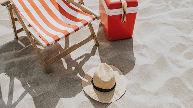Un chapeau de paille, une glacière et une chaise de plage sur une plage de sable fin. 
