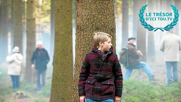 Un jeune garçon caché derrière un arbre pendant que des hommes fouillent la forêt.