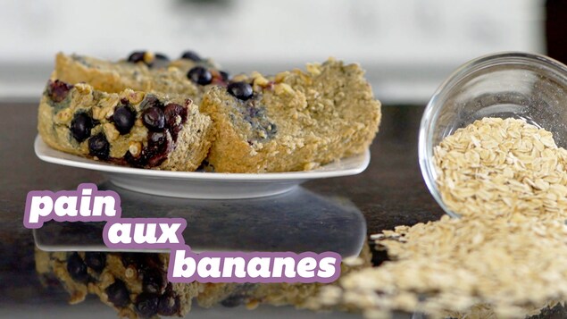 Le titre pain aux bananes est écrit en blanc avec un contour mauve en-bas à gauche de l'image. Trois tranches de pain aux bananes avec des bleuets agissent en fond d'écran. Un bol d'avoine renversé est situé à droite de l'image.