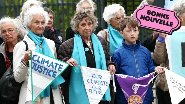 Des femmes âgées de 70 ans et un jeune garçon tiennent une banderole avec des messages de justice climatique à côté du logo Bonne nouvelle de MAJ.