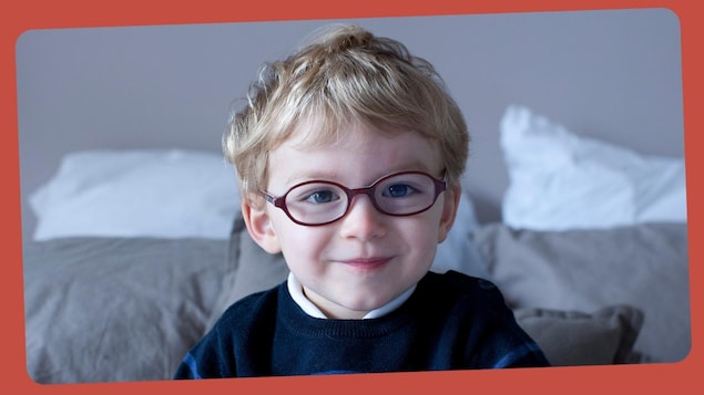 Il porte des lunettes, un pull rayé et un jean. Il est assis près d'un lit. 