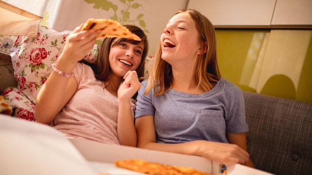 Deux amies ados rient en mangeant de la pizza, assises dans un canapé