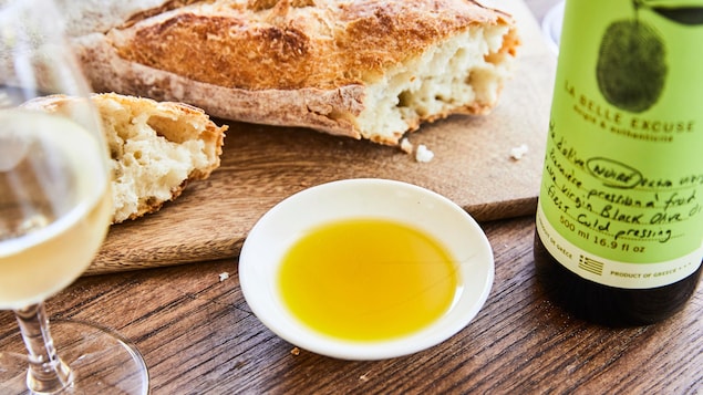 Un petit ramequin rempli d'huile d'olive extra-vierge, entouré d'une bouteille d'huile, d'un verre de vin blanc et d'une miche de pain baguette sur une planche en bois.