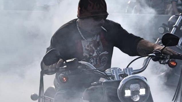 Un homme avec des lunettes fumées, une tuque ornée de flames et une cigarette entre les lèvres conduit une motocyclette, entouré de fumée.