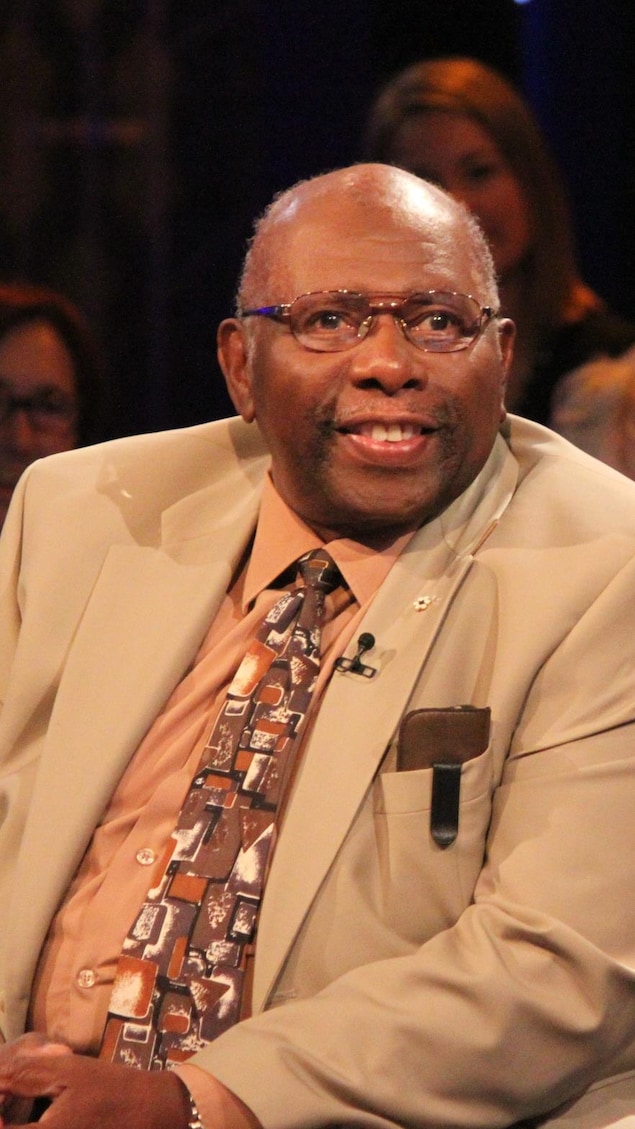 Oliver Jones souriant assis sur un fauteuil de plateau de télévision.