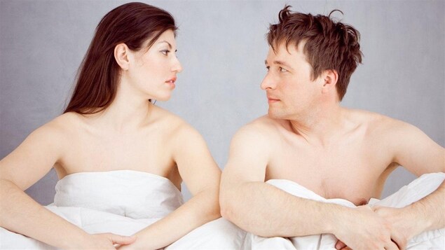 Un couple, assis dans un lit, se regarde.
