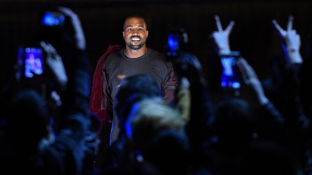 Kanye West sourit devant le public qui le prend en photo avec des téléphones intelligents.