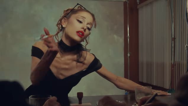 Ariana Grande dans le vidéoclip pour la chanson Petal. Elle porte une robe noire qui montre son sternum et tient une cigarette entre ses doigts.