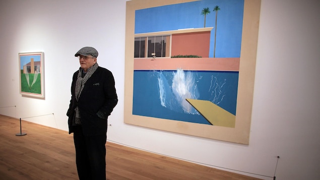 David Hockney debout devant un de ses tableaux dans une galerie d'art.