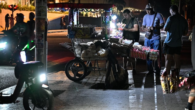 Des clients achètent des denrées à un vendeur de rue, dont l'étal est éclairé par une lampe à batteries.