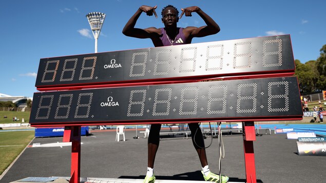 Près d'une piste d'athlétisme, un sprinteur victorieux pointe un chronomètre géant.