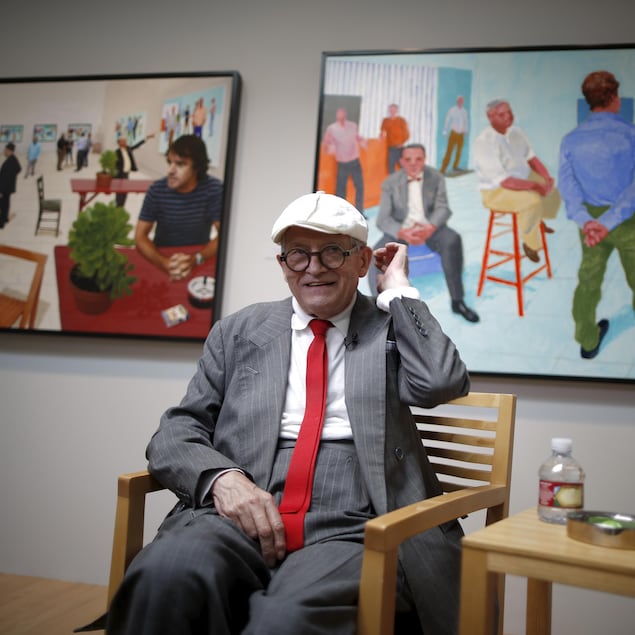 David Hockney souriant, assis sur une chaise de bois, devant deux tableaux.