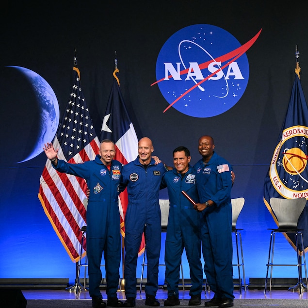 Quatre hommes en combinaison bleue sur une scène, devant des drapeaux des États-Unis, du Texas, de l'Agence spatiale européenne et de la NASA