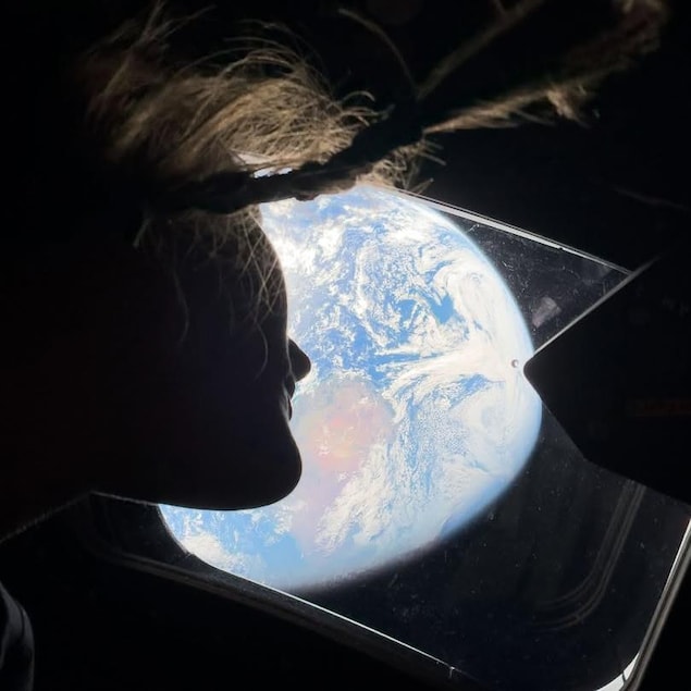 Contre-plongée d'une femme à contrejour qui regarde la terre à travers le hublot d'une capsule spatiale.