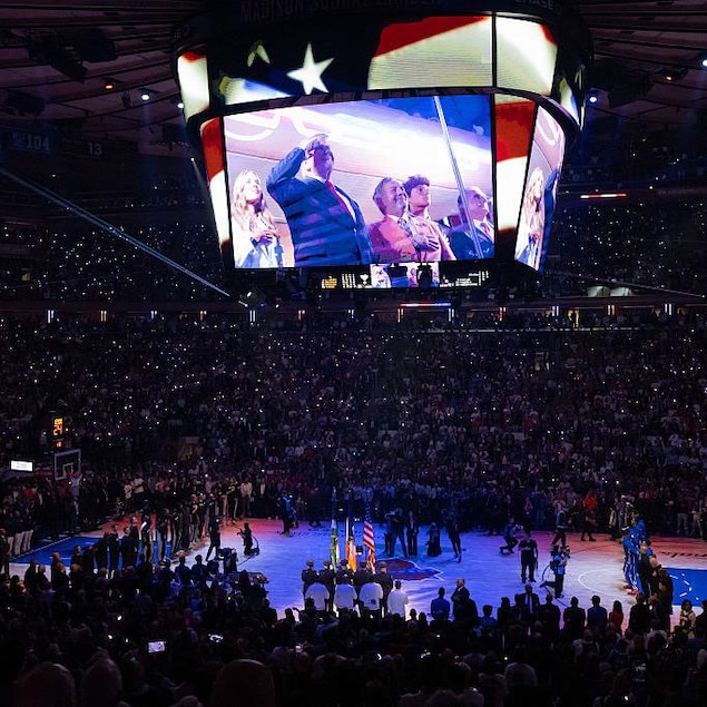 Pendant l'hymne national d'un match de basketball, un écran géant au-dessus du court et de la foule montre Donald Trump qui fait le salut militaire, aux côtés d'autres personnes, souriantes.