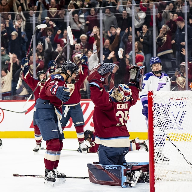 Sur une patinoire, des joueuses de hockey célèbrent la victoire, devant une foule en liesse.