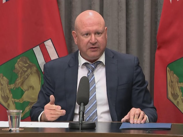 Un homme chauve en complet lors d'une conférence de presse. Il est assis devant des drapeaux du Manitoba.