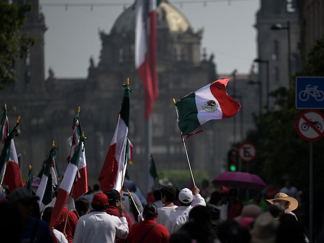 Des gens de dos marchent dans la rue en tenant des drapeaux mexicains.