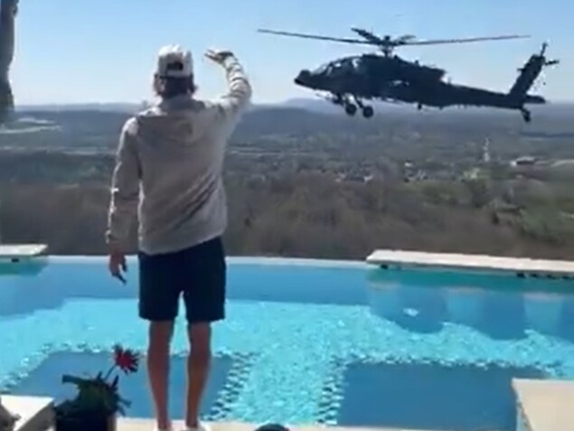 Dans une résidence située en hauteur, un homme debout près d'une piscine creusée envoie la main à un hélicoptère qui vole à proximité.