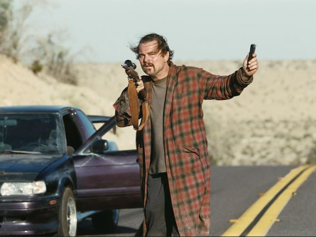 Leonardo DiCaprio dans une scène de film où il marche en robe de chambre sur une route en tenant une carabine.