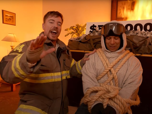MrBeast et un cascadeur ligoté, qui porte de lunettes de ski sur sa tête. Ils se tiennent devant un feu et une table sur laquelle sont déposés des sacs d'argent.