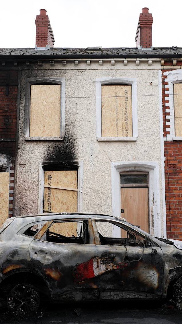 Une voiture calcinée devant des maisons incendiées dont les fenêtres et les portes sont placardées.