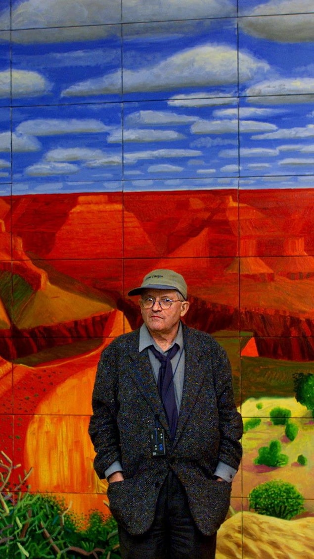 David Hockney debout, les mains dans les poches, devant une de ses toiles représentant le Grand Canyon.
