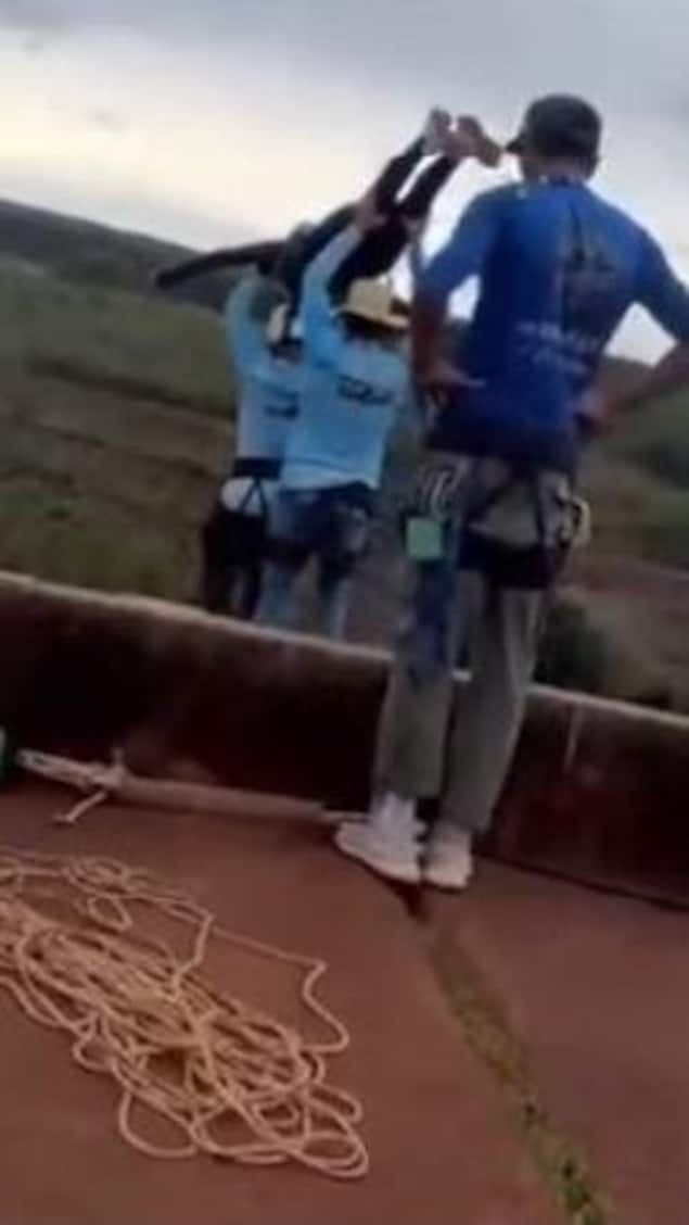 Deux hommes portent à bout de bras une femme munie d'un casque. Umn autre les regarde, une corde à ses pieds.