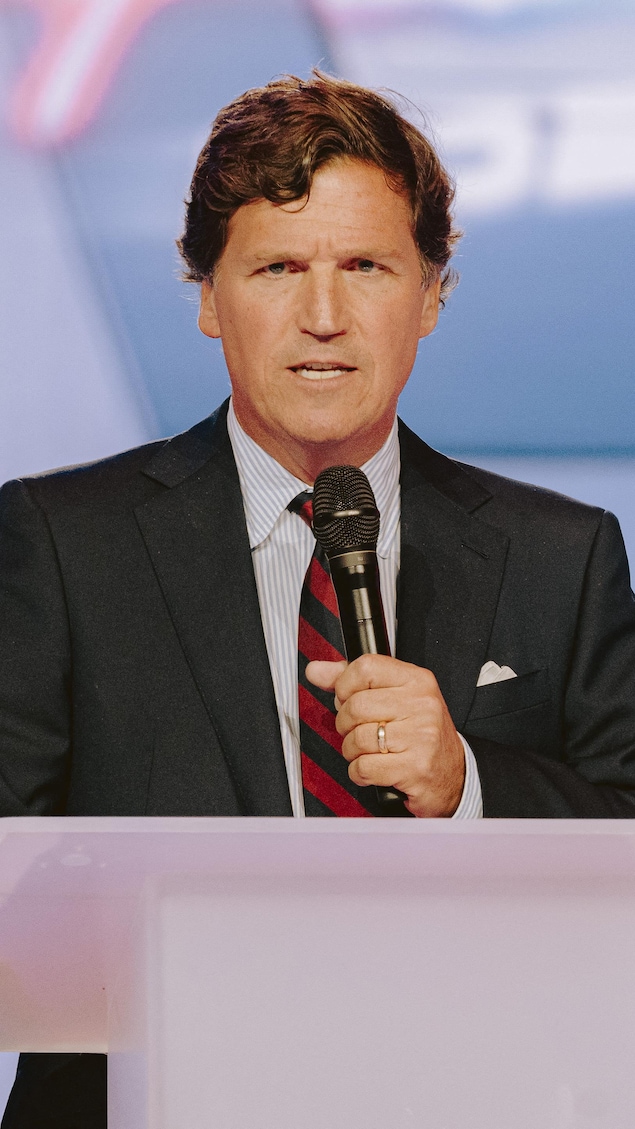 Sur une scène, derrière un lutrin, Tucker Carlson parle dans un micro.