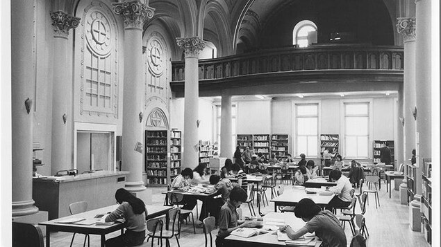 Vue en plan large d'une grande salle avec des tables où des étudiants sont au travail. Des étagères avec des livres meublent certains des murs qui les entourent. Vue en plan large d'une grande salle avec des tables où des étudiants sont au travail. Des étagères avec des livres meublent certains des murs qui les entourent.