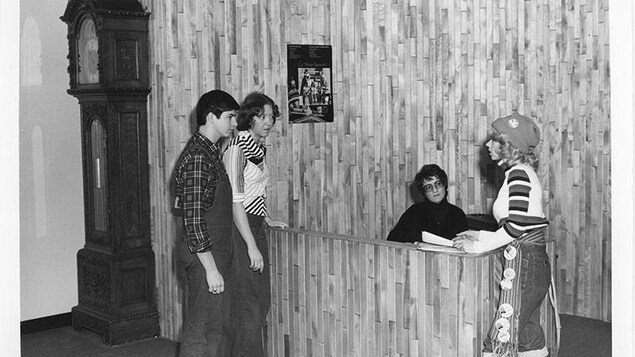 Quatre personnes, trois étudiants et une réceptionniste sont autour de la réception. Les murs sont faits de lattes de bois. À la gauche, il y a une grande horloge grand-père. Quatre personnes, trois étudiants et une réceptionniste sont autour de la réception. Les murs sont faits de lattes de bois. À la gauche, il y a une grande horloge grand-père.