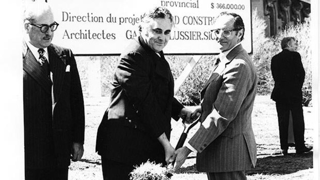 À l'avant-plan, trois hommes sont debout et deux d'entre eux tiennent une pelle en regardant le photographe et en souriant, devant un édifice et une grande affiche qui détaille les éléments du projet de construction. À l'avant-plan, trois hommes sont debout et deux d'entre eux tiennent une pelle en regardant le photographe et en souriant, devant un édifice et une grande affiche qui détaille les éléments du projet de construction.